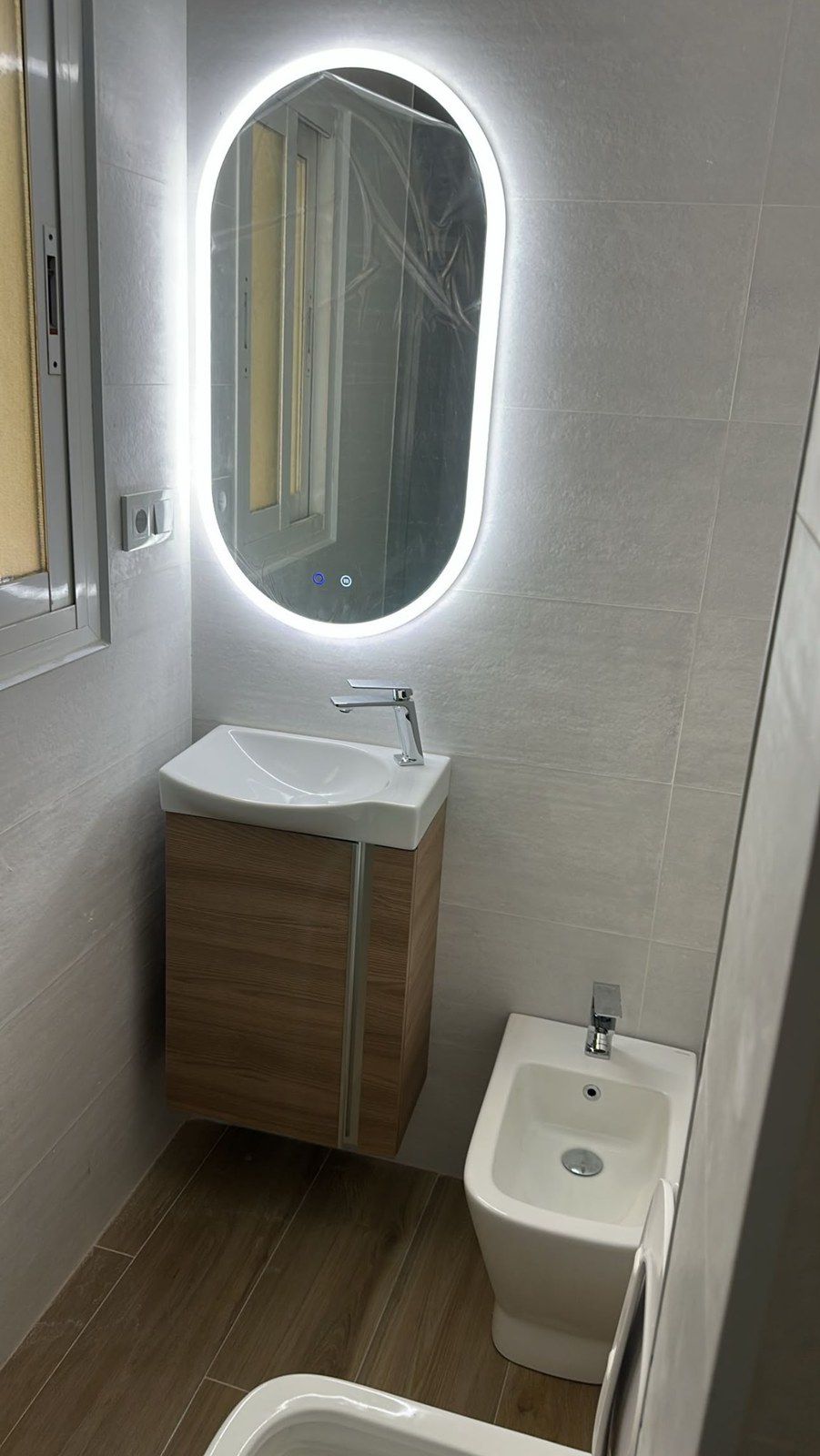 Baño reformado con espejo ovalado retroiluminado y mueble de madera