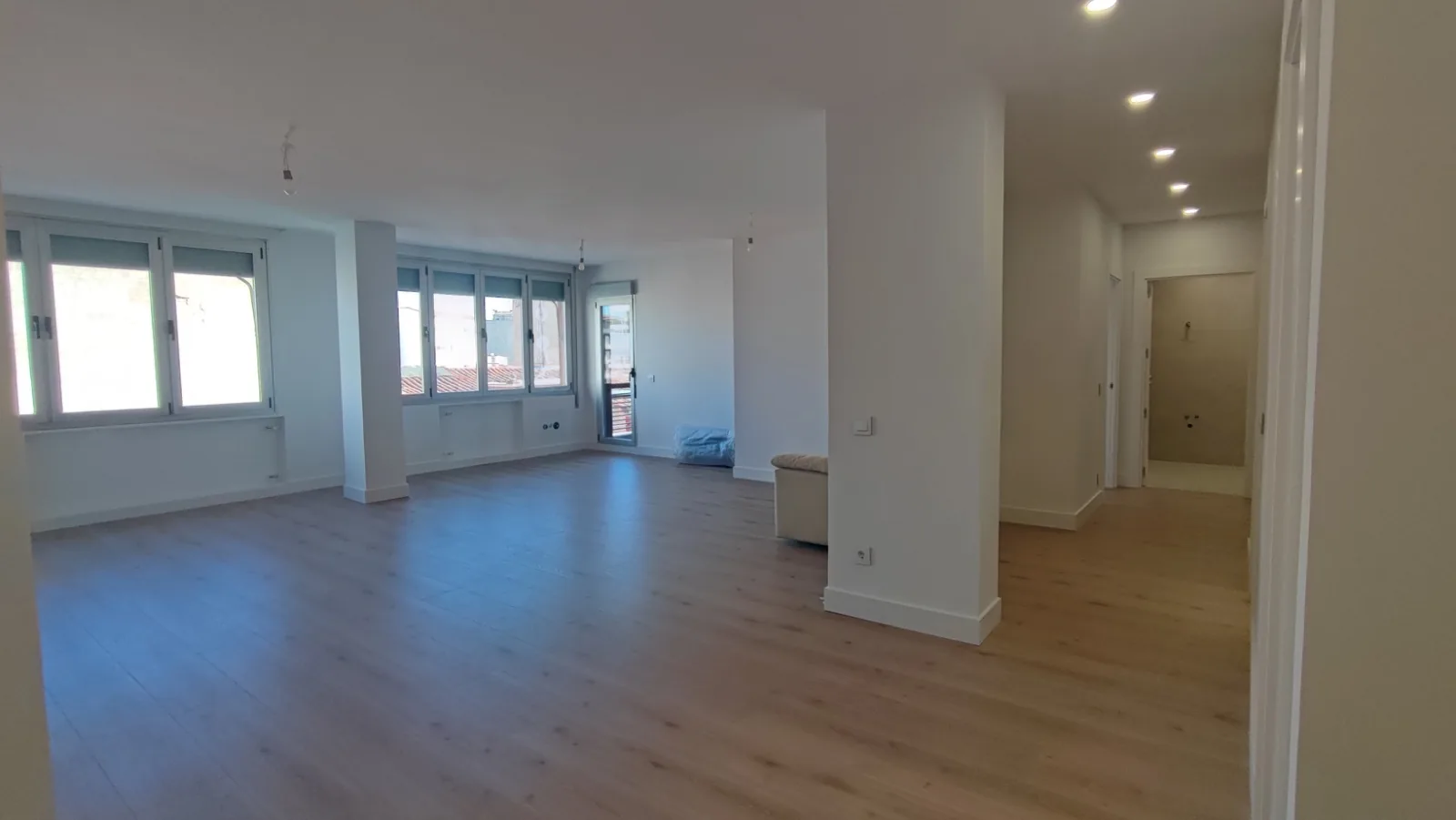 Salón diáfano de piso reformado con suelo laminado, focos y ventanas al fondo