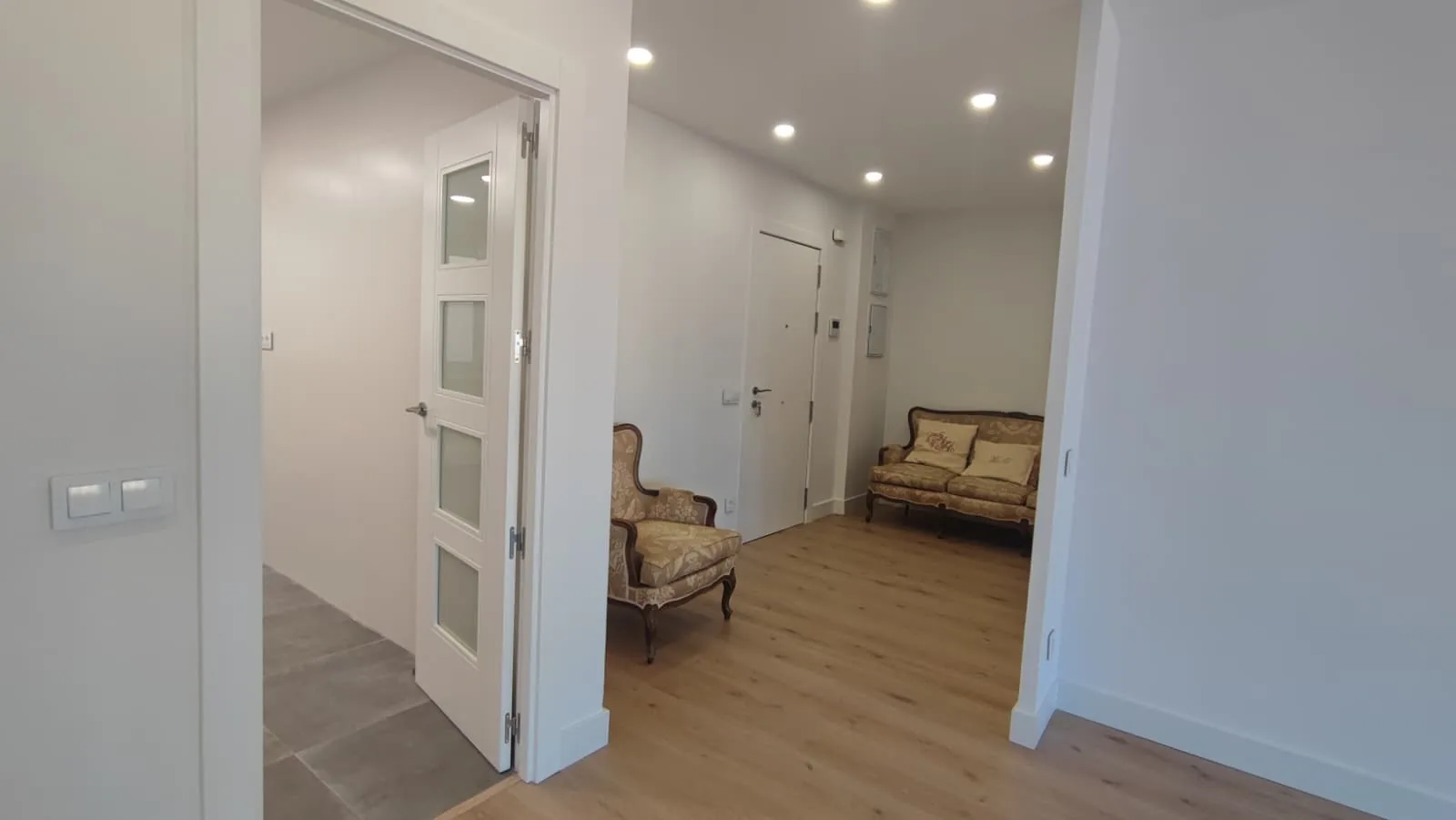 Recibidor de vivienda reformada con puerta de cristal, focos y sillones clásicos