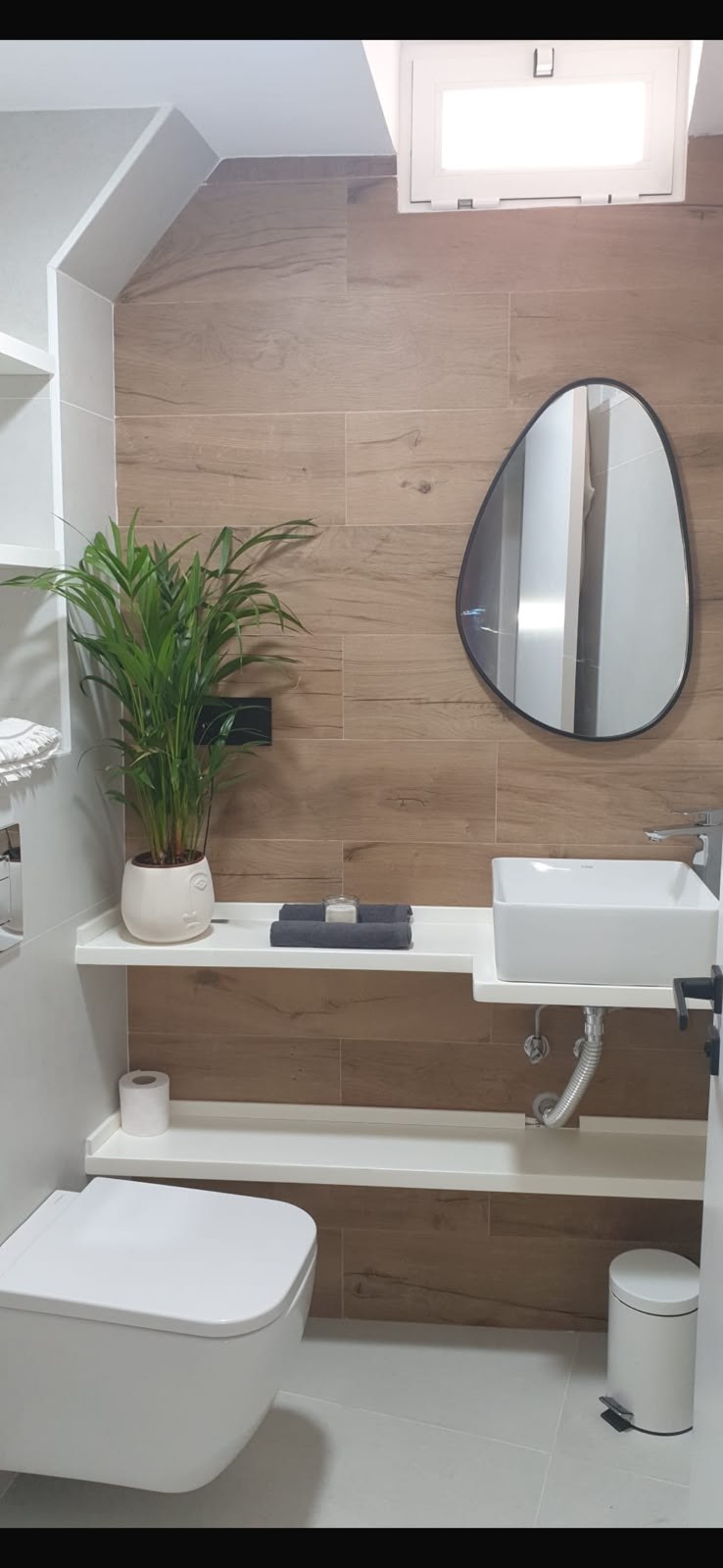 Baño con revestimiento efecto madera, espejo orgánico e inodoro suspendido