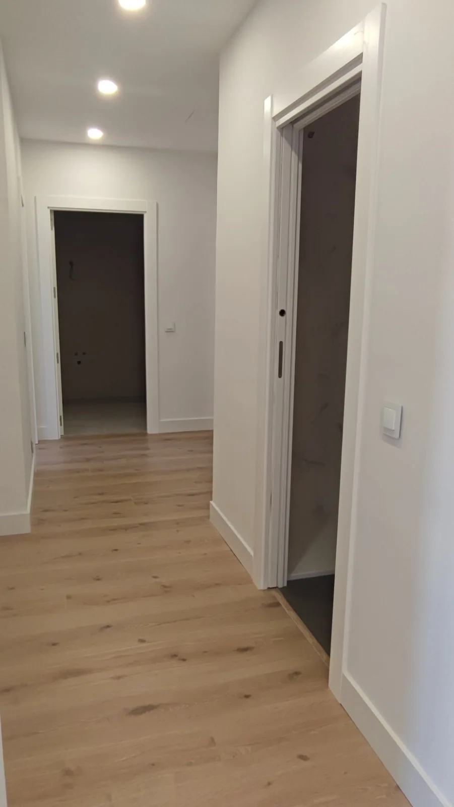 Pasillo de un piso reformado con suelo de madera clara, focos empotrados y puertas blancas