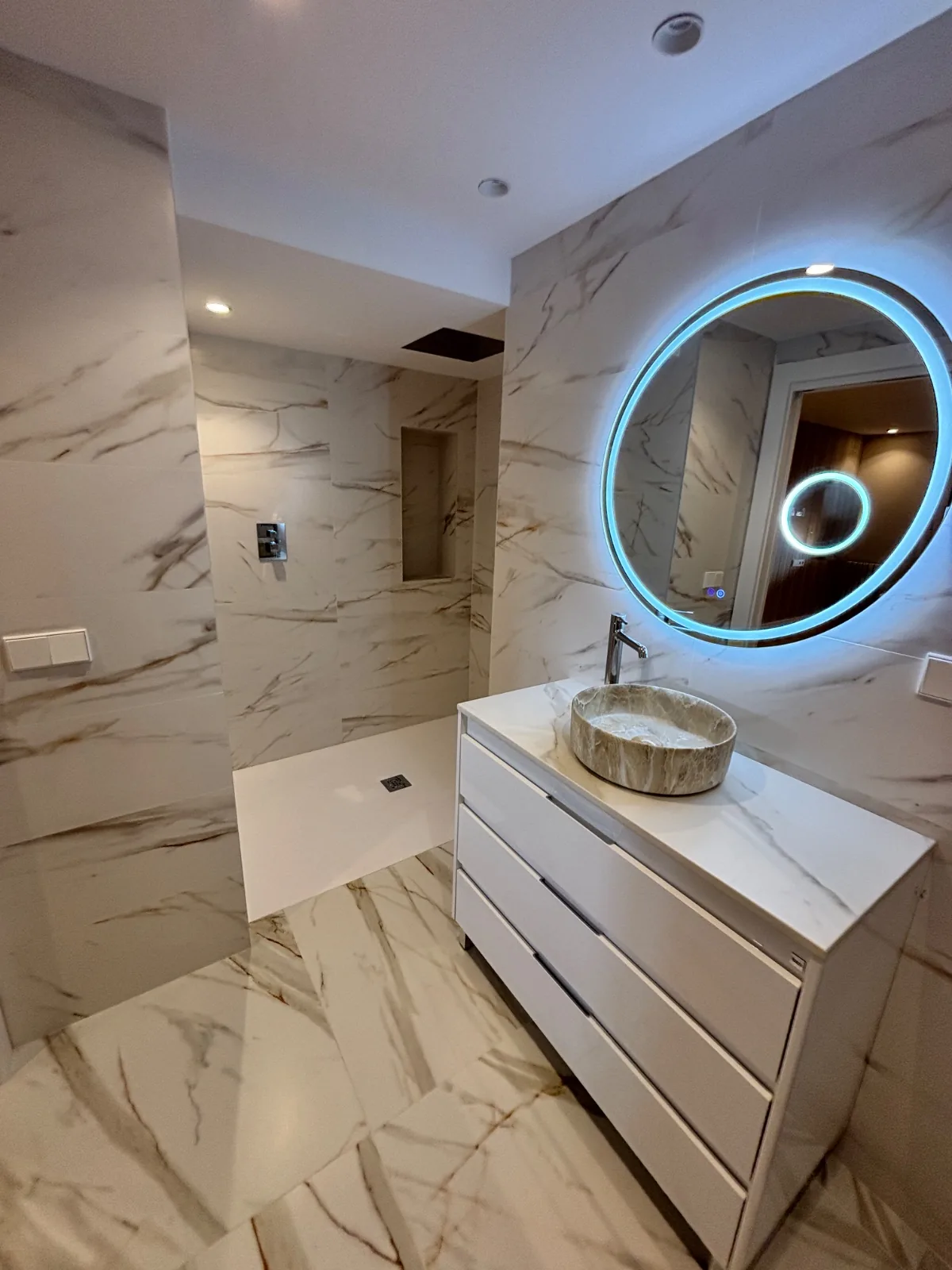 Baño con suelo y paredes efecto mármol, mueble con lavabo sobre encimera y ducha de obra